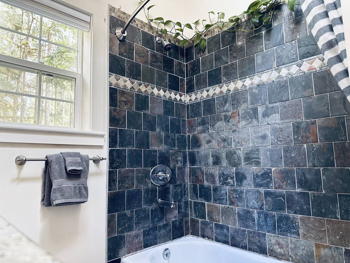 Primary bath tub and shower in dark slate tile with a stone diamond border, a window onto the forest and grey towels
