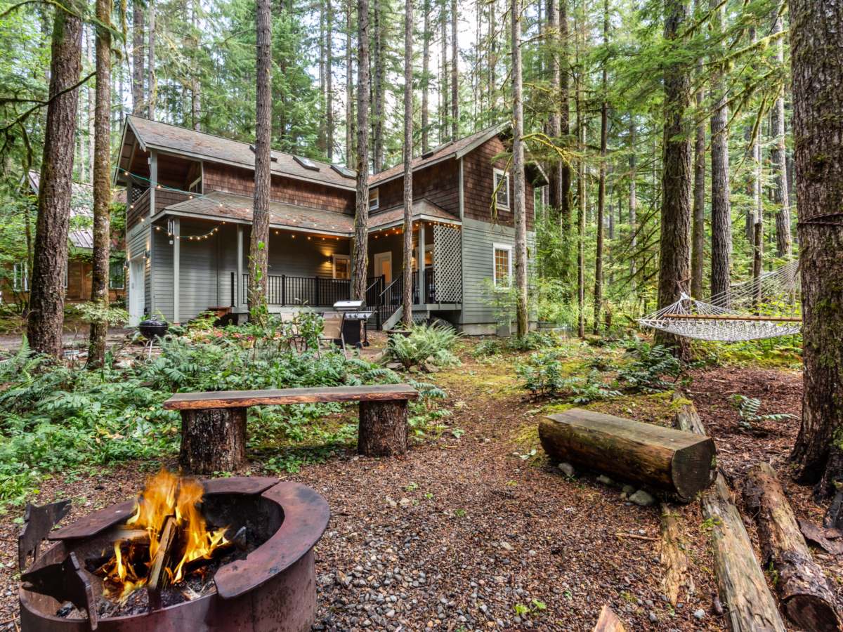 Two-story cedar-shingle cabin among tall firs, with string lights on the covered porch, a gas grill, a rope hammock and a lit fire pit ringed by log benches in the foreground