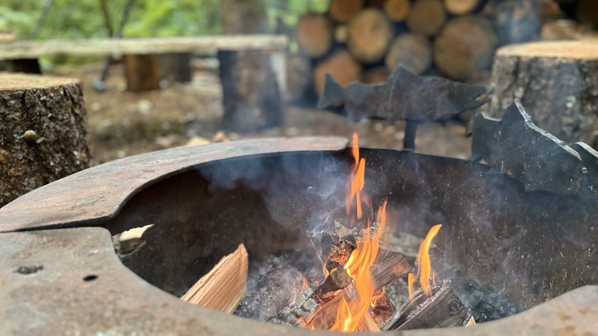 Flames in the steel fire pit with a leaping-salmon cutout, log-round stools and stacked firewood behind