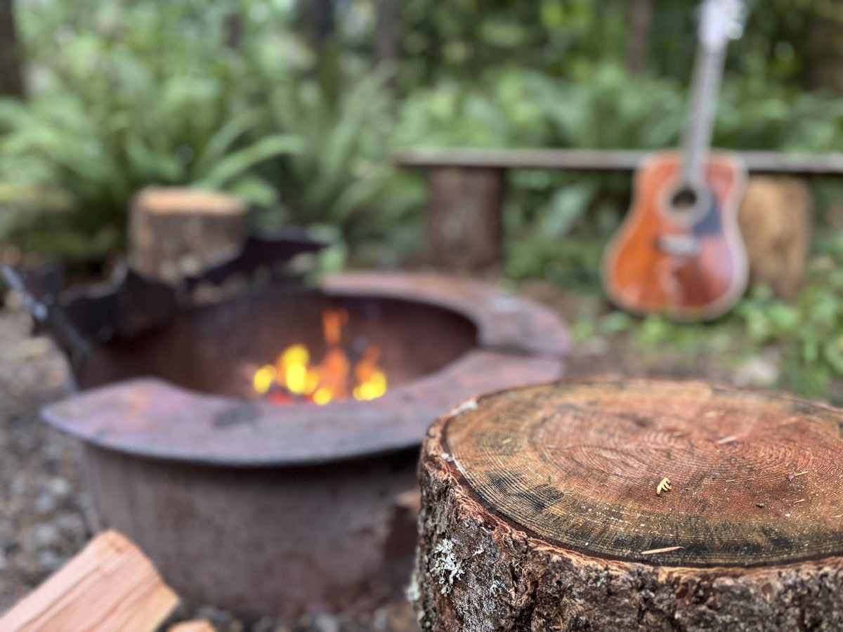 A glowing fire in the steel fire pit, a round log stool in front and an acoustic guitar leaning on a log bench