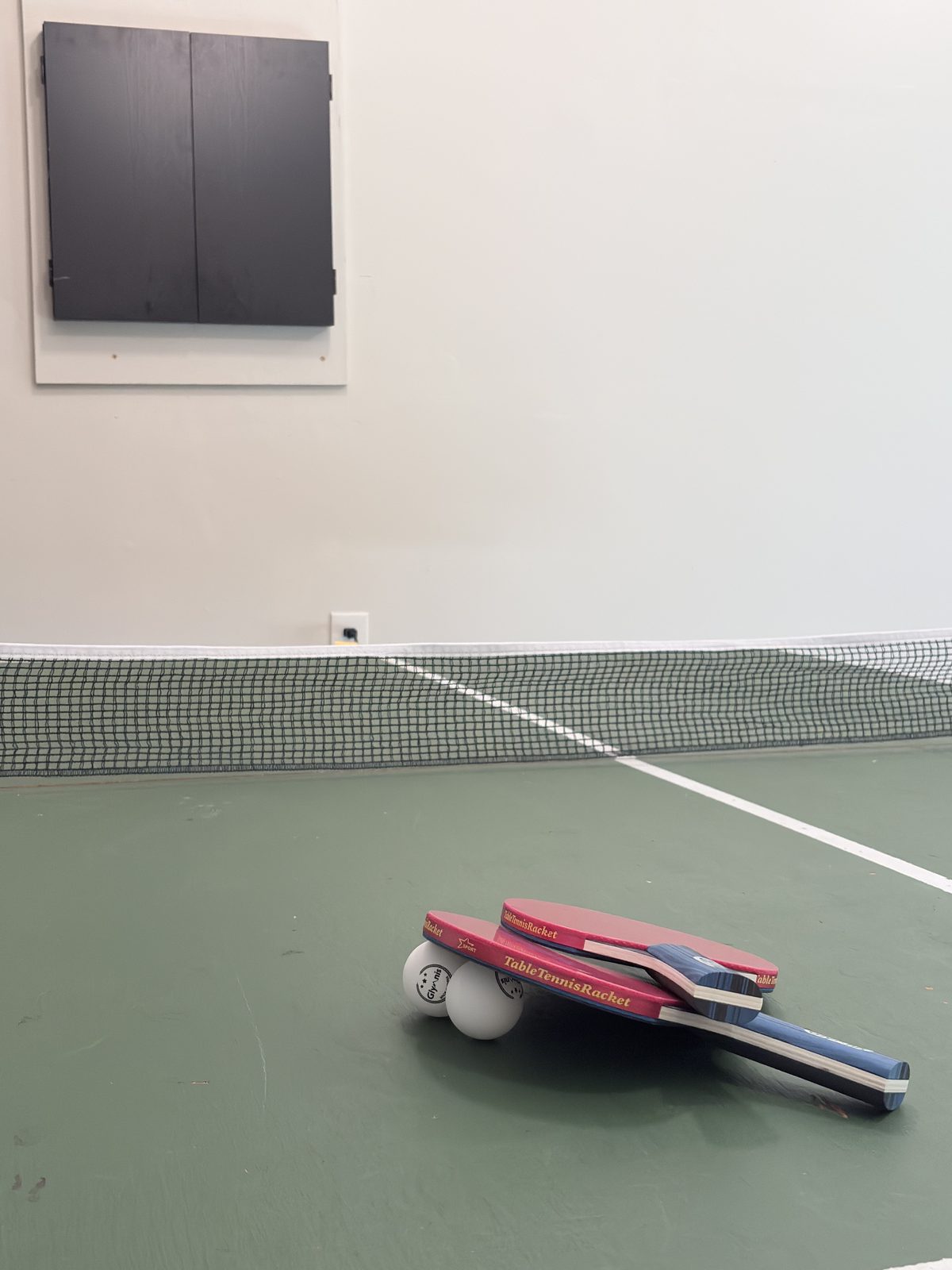 Two ping pong paddles and balls on the green table in the game room, with a closed dartboard cabinet on the wall