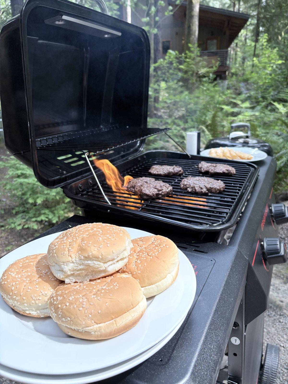 Burgers on the gas grill in the yard beside a plate of sesame buns, with the charcoal kettle and forest behind