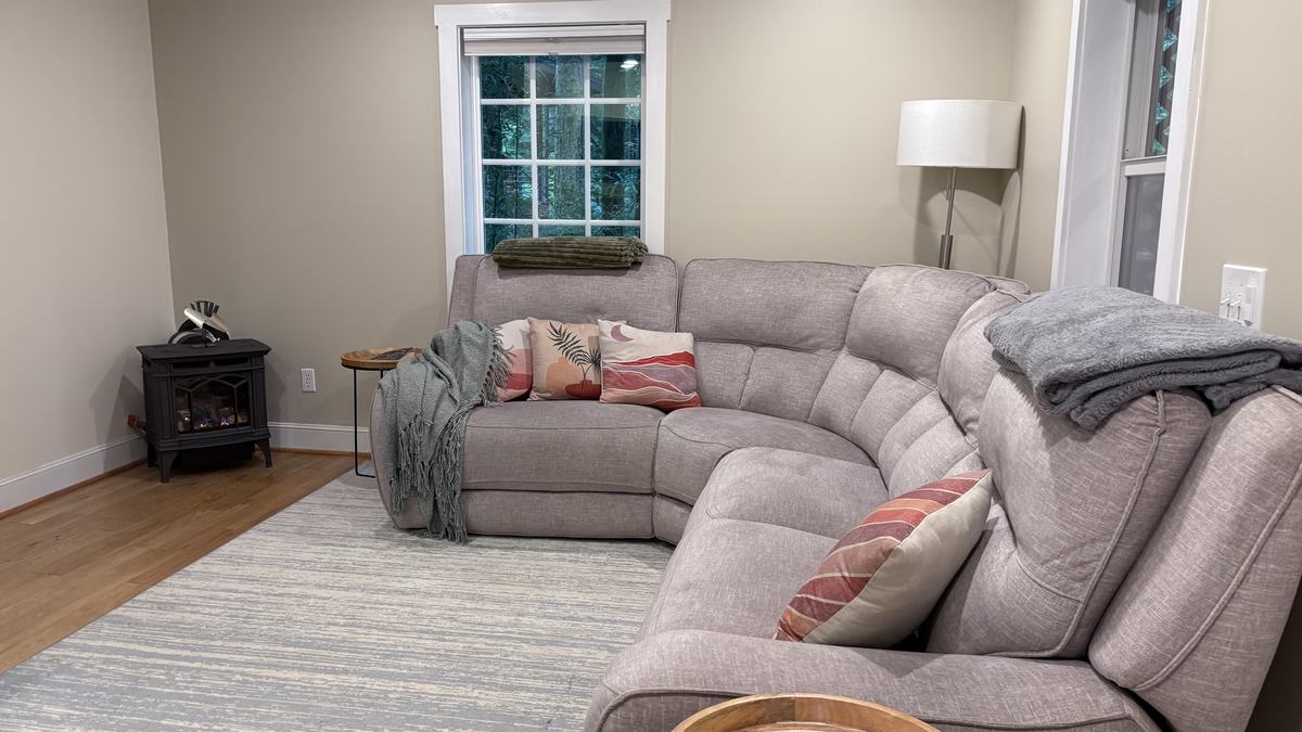 The main living room: a large grey sectional with throws and patterned pillows, a floor lamp, a window onto the forest and a black gas stove in the corner