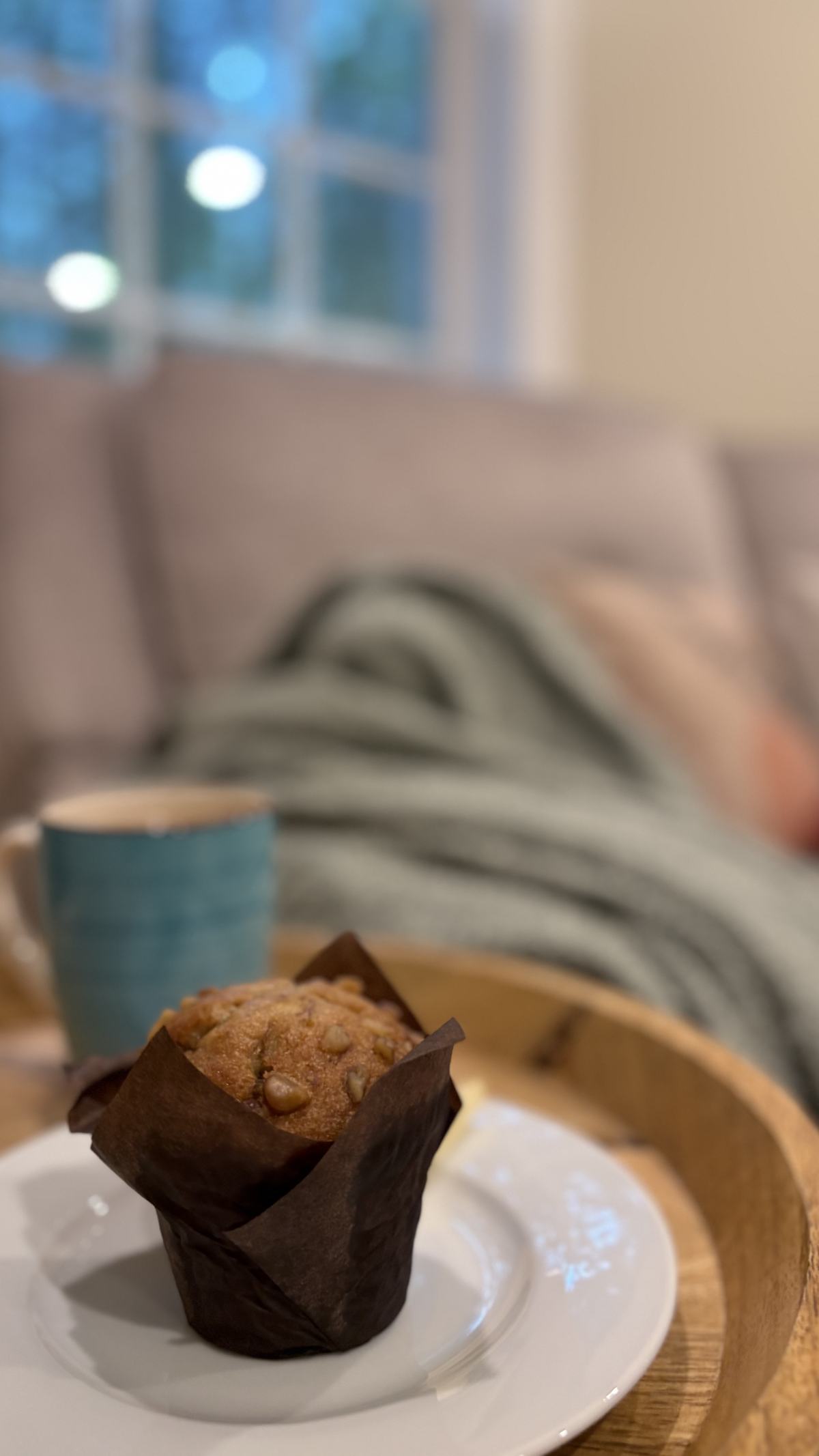 A walnut muffin on a white plate and a teal mug on a wooden tray, with a knit throw on the sectional behind