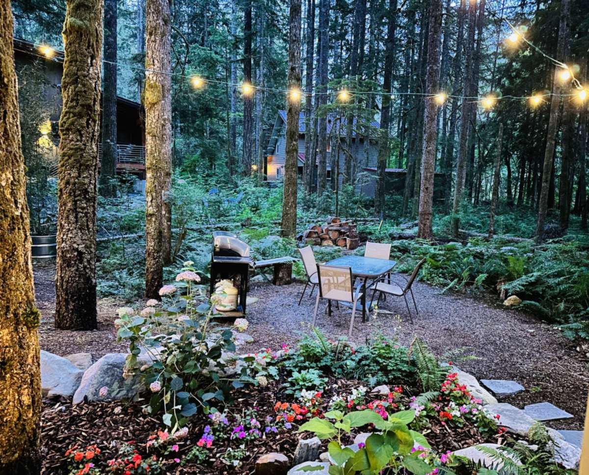 The yard at dusk under string lights: a gas grill and a patio table with four chairs on gravel, ringed by ferns, flower beds and tall moss-covered firs