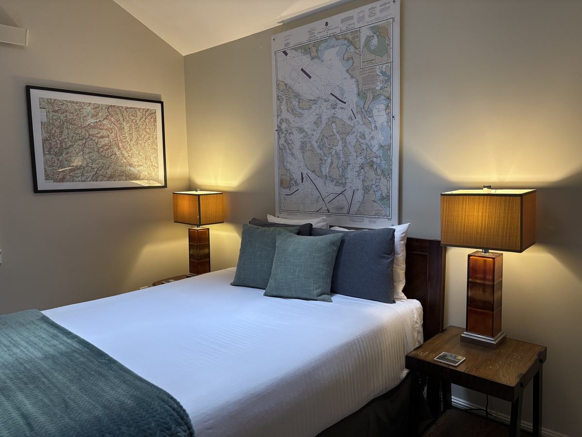 The queen bedroom: white bedding with teal and blue pillows, a teal throw, amber table lamps on wood nightstands, a large nautical chart above the bed and a framed relief map on the side wall