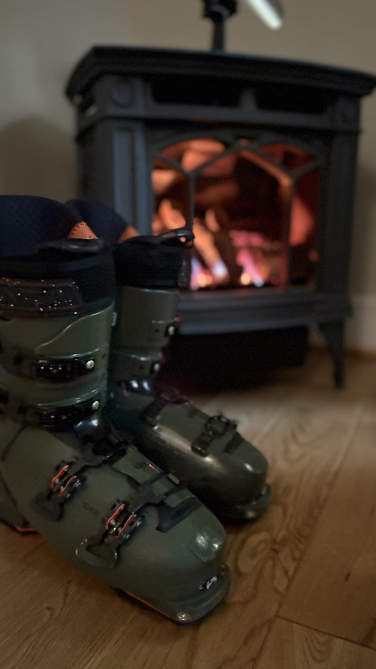 Ski boots drying on the wood floor in front of the gas stove with its flames lit
