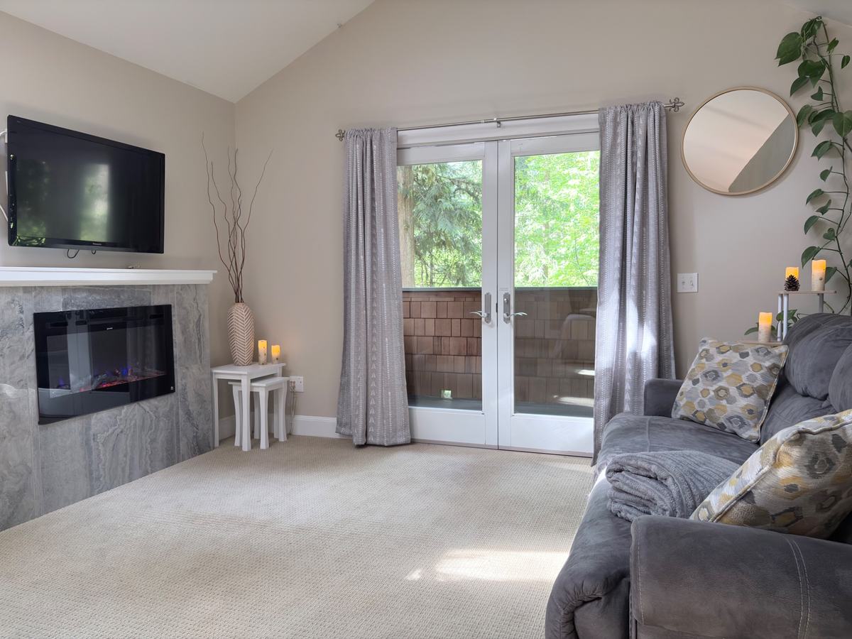 The king suite sitting area: a lit electric fireplace in grey stone tile, a wall-mounted TV, and French doors opening to the cedar-shingled balcony and trees