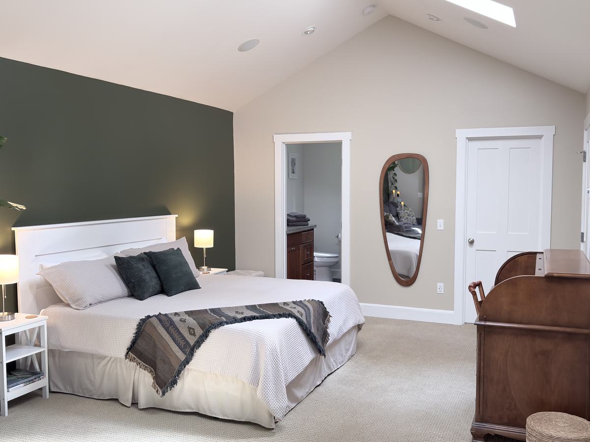 The king bed with a white headboard and dark green pillows against a forest-green accent wall, with the door to the en suite bath, a tall wood-framed mirror and the roll-top desk