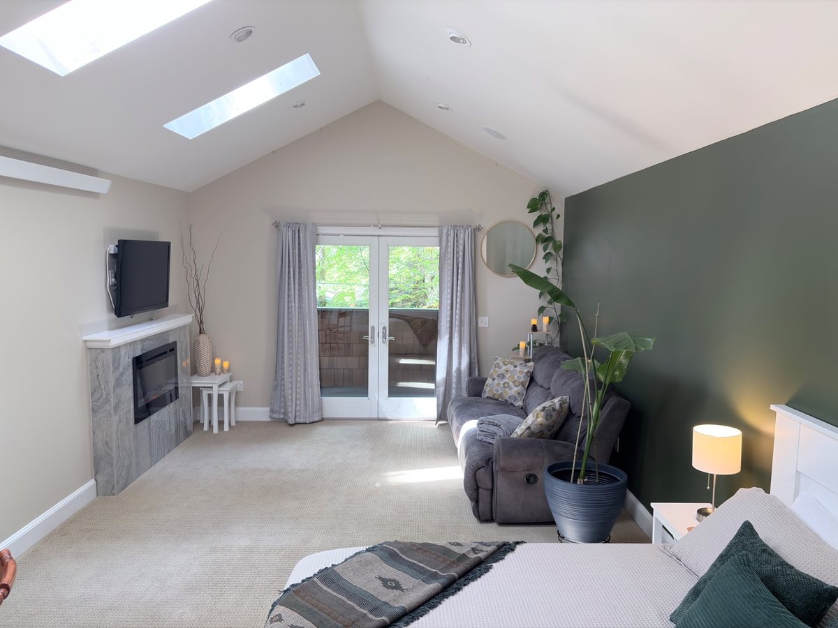 The king suite under a vaulted ceiling with two skylights: a dark green accent wall behind the bed, a reclining sofa, an electric fireplace with a TV above, and French doors to the balcony